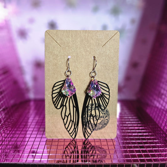 Image shows a brown card with stainless steel earring wires that have black metal cutout fairy wings and green and purple baroque-style crystals hanging from the wires.