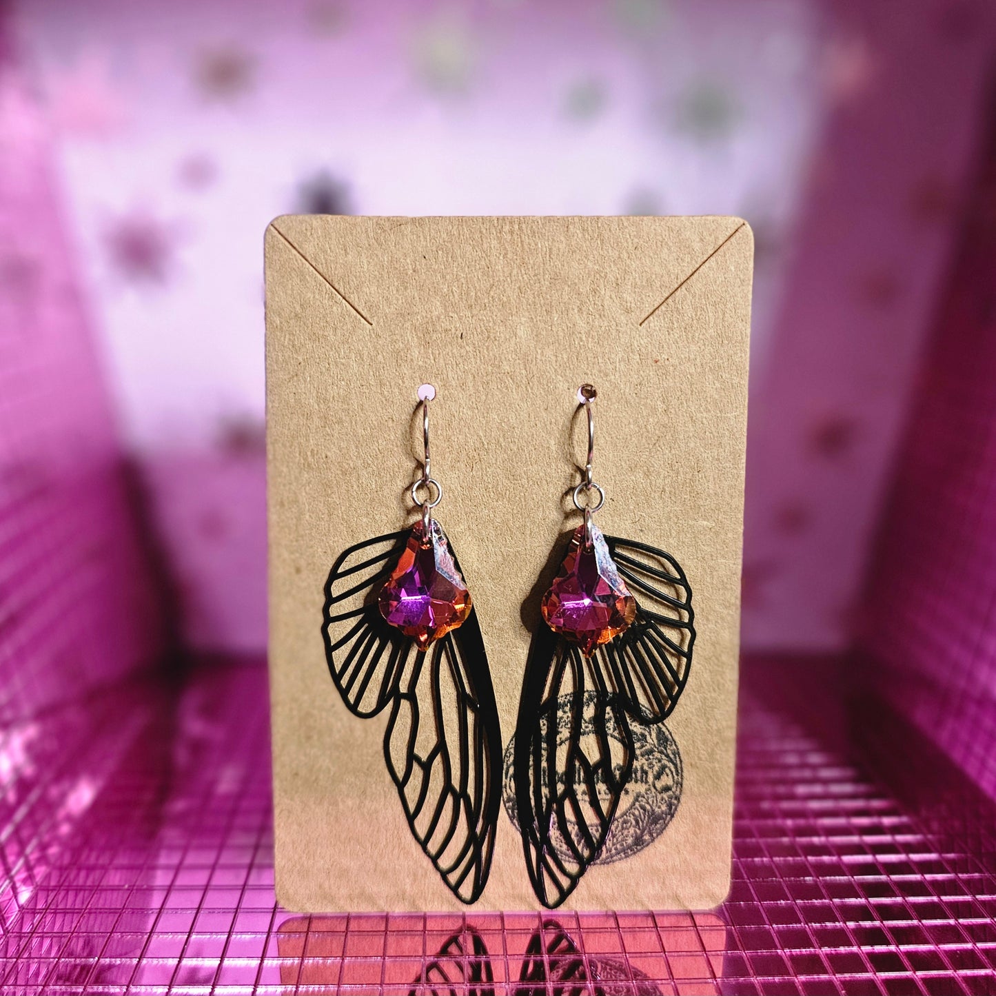 Image shows a brown card with stainless steel earring wires that have black metal cutout fairy wings and red and purple baroque-style crystals hanging from the wires.