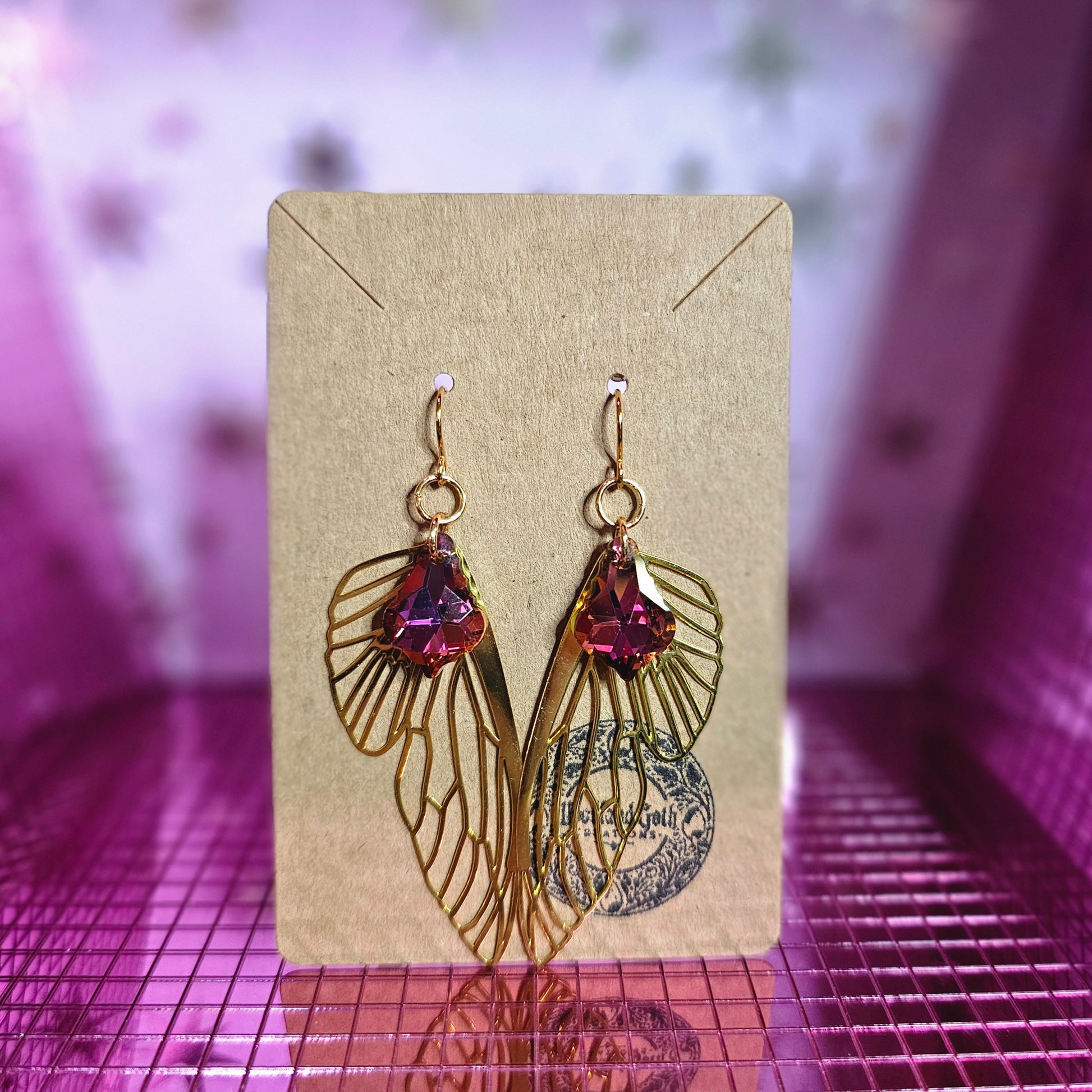 Image shows a brown card with 14k gold plated stainless steel earring wires that have golden metal cutout fairy wings and red and purple baroque-style crystals hanging from the wires.